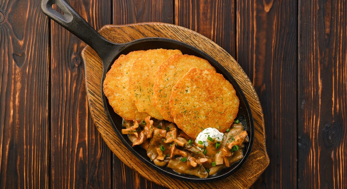4 Kartoffelpuffer mit Rahm-Pfifferlingen in einer gusseisernen Pfanne, die auf einem Brett steht, Draufsicht.