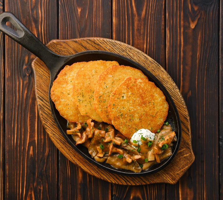 4 Kartoffelpuffer mit Rahm-Pfifferlingen in einer gusseisernen Pfanne, die auf einem Brett steht, Draufsicht. | © Adobe Stock/  boomeart