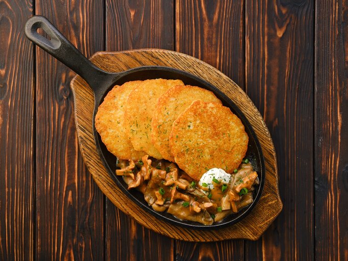 4 Kartoffelpuffer mit Rahm-Pfifferlingen in einer gusseisernen Pfanne, die auf einem Brett steht, Draufsicht. | © Adobe Stock/  boomeart