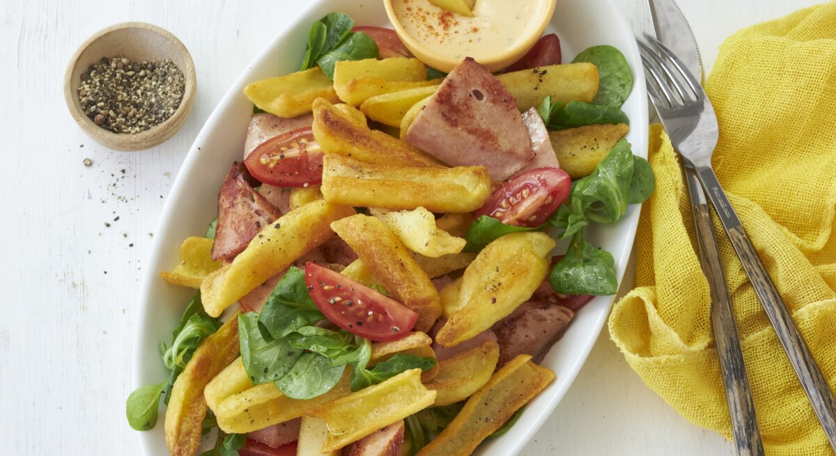 Draufsicht: Heller Untergrund auf dem ein ovaler Teller mit bayerischen Pommes mit Leberkäse und Obazda-Dip steht. Garniert mit ein paar Blättern Feldsalat und Tomatenspalten. Rechts daneben eine gelbe Stoffservierte und rustikales Holzbesteck.