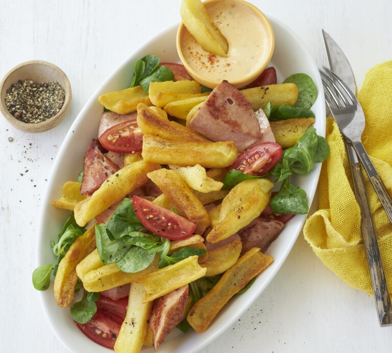 Draufsicht: Heller Untergrund auf dem ein ovaler Teller mit bayerischen Pommes mit Leberkäse und Obazda-Dip steht. Garniert mit ein paar Blättern Feldsalat und Tomatenspalten. Rechts daneben eine gelbe Stoffservierte und rustikales Holzbesteck. | © StockFood / StockFood Studios / Brachat, Oliver