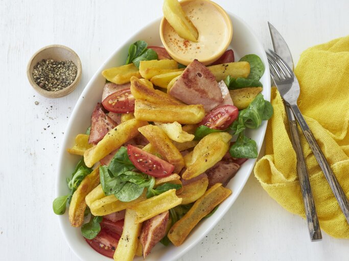 Draufsicht: Heller Untergrund auf dem ein ovaler Teller mit bayerischen Pommes mit Leberkäse und Obazda-Dip steht. Garniert mit ein paar Blättern Feldsalat und Tomatenspalten. Rechts daneben eine gelbe Stoffservierte und rustikales Holzbesteck. | © StockFood / StockFood Studios / Brachat, Oliver
