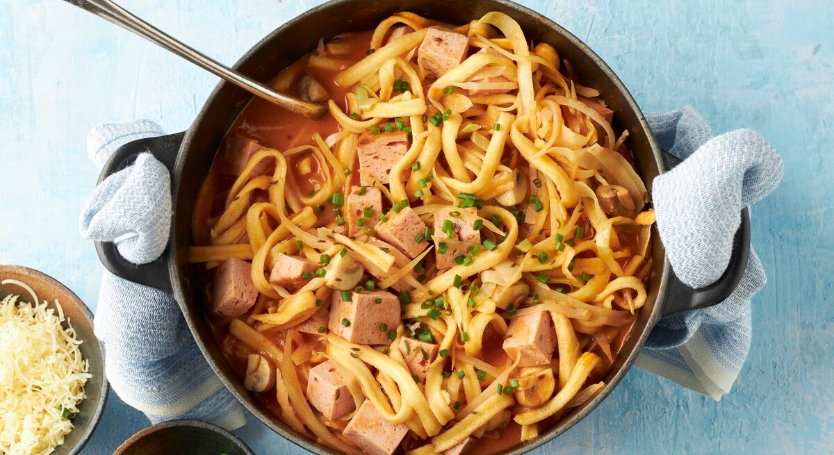 In einem Topf ist ein bayerischer Spätzle-Topf mit Leberkäse, Champignons und Kohl angerichtet. Daneben steht geriebener Käse.