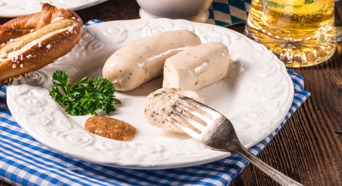 Ein Teller mit veganen Weiswürsten, süßem Senf und einer Brezel, daneben ein Glas Bier.