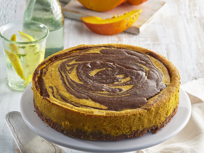 Ein marmorierter Kürbis-Käsekuchen steht auf einem Kuchenpodest. Der Untergrund ist aus hellem Holz. Um das Podest herum liegt ein Geschirrtuch, ein Brett mit Kürbisstückchen, ein Glas und eine Flasche mit Wasser und ein Kuchenheber.  | © Stockfood / Profimedia