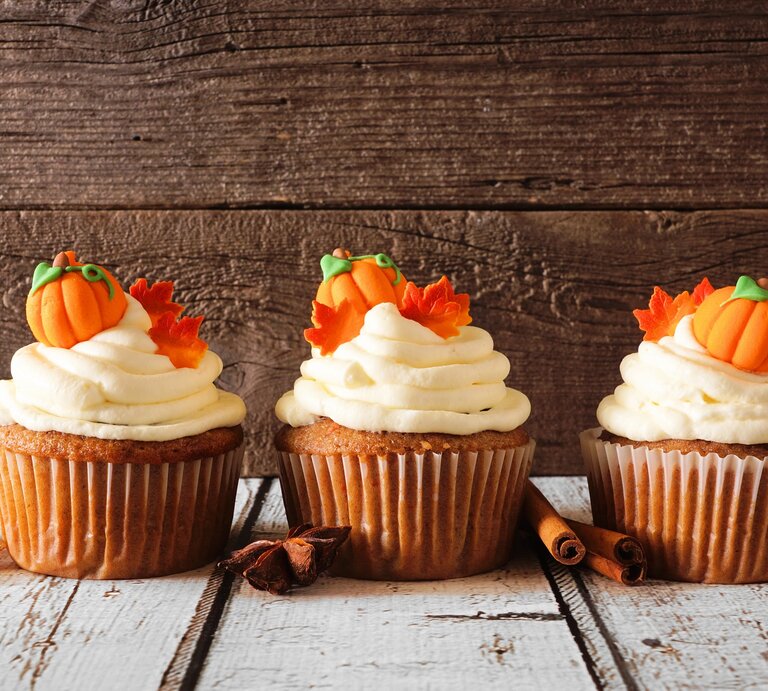 Drei Kürbis-Cupcakes verziert mit kleinen Kürbissen und Ahornblättern aus Marzipan, daneben Zimtstangen und Anissterne. | © Adobe Stock/ Jenifoto