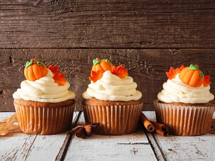 Drei Kürbis-Cupcakes verziert mit kleinen Kürbissen und Ahornblättern aus Marzipan, daneben Zimtstangen und Anissterne. | © Adobe Stock/ Jenifoto