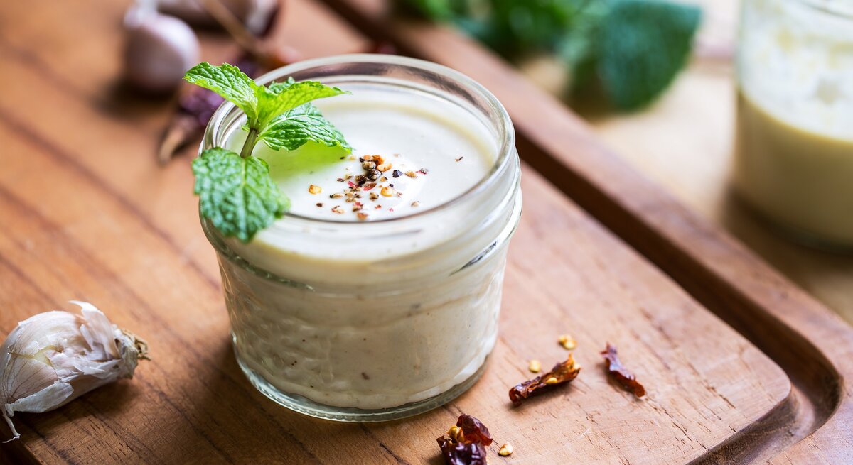 Ein Glas weißes Knoblauch-Zitronen-Dressing mit Minze und Pfeffer dekoriert, drumherum frische Zutaten.