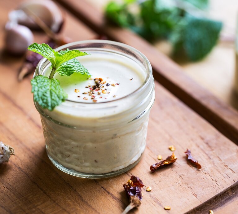 Ein Glas weißes Knoblauch-Zitronen-Dressing mit Minze und Pfeffer dekoriert, drumherum frische Zutaten. | © Adobe Stock/ vanillaechoes
