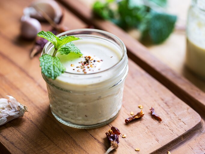 Ein Glas weißes Knoblauch-Zitronen-Dressing mit Minze und Pfeffer dekoriert, drumherum frische Zutaten. | © Adobe Stock/ vanillaechoes
