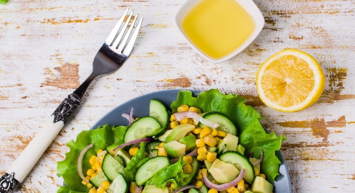 Schale mit Mango-Dressing neben einem Teller Salat mit Gurke und Mais auf hölzernem Untergrund. 