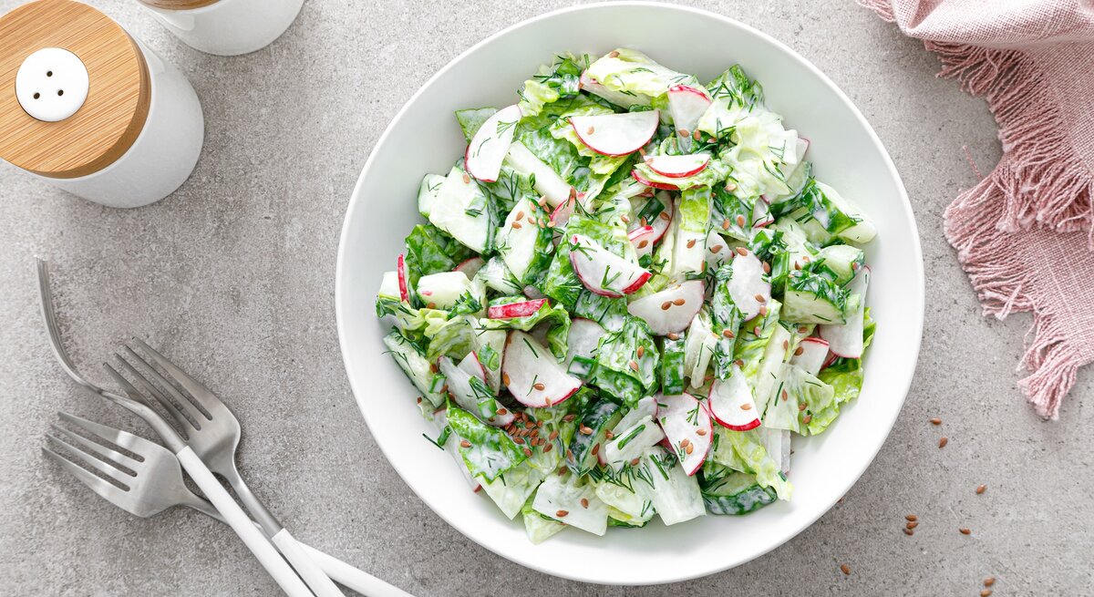 Draufsicht: Grauer Untergrund, eine Schüssel mit einem Gurken-Radieschen-Salat. Daneben stehen Salz- und Pefferstreuer, ein Besteck sowie ein Stoffdecke.
