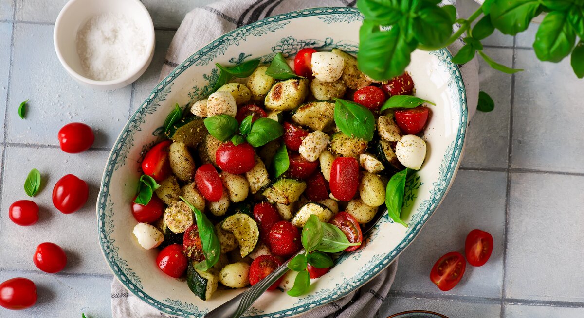 Gnocchi-Salat Caprese mit Tomaten, Zucchini und Mozzarella in kitschiger Schale auf weißen Fliesen. Daneben Basilikum, Salz, kirschtomaten und Olivenöl.
