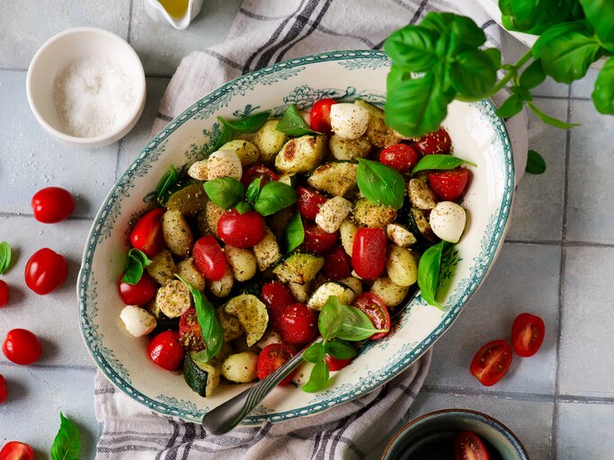 Gnocchi-Salat Caprese mit Tomaten, Zucchini und Mozzarella in kitschiger Schale auf weißen Fliesen. Daneben Basilikum, Salz, kirschtomaten und Olivenöl. | © AdobeStock/zoryanchik