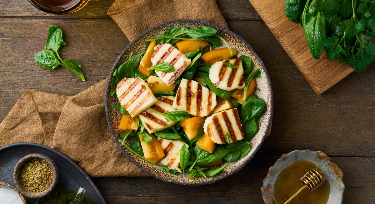 Untergund aus Holz. Darauf ein Teller  Gegrillter Halloumi-Salat mit orangen Tomaten. Drum herum liegen Spinatblätter und grobe Leinentücher