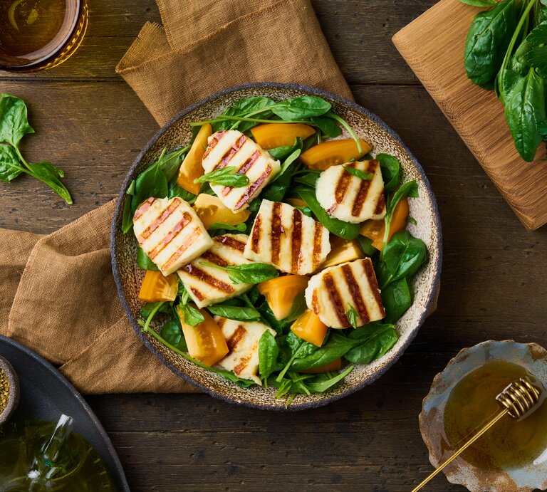 Untergund aus Holz. Darauf ein Teller  Gegrillter Halloumi-Salat mit orangen Tomaten. Drum herum liegen Spinatblätter und grobe Leinentücher | © Shutterstock / Nata Bene