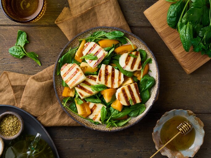 Untergund aus Holz. Darauf ein Teller  Gegrillter Halloumi-Salat mit orangen Tomaten. Drum herum liegen Spinatblätter und grobe Leinentücher | © Shutterstock / Nata Bene
