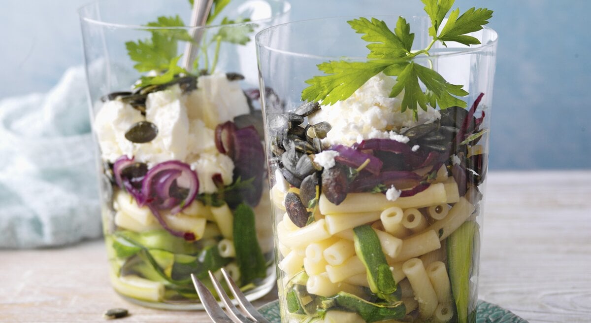 Zucchini-Makkaronisalat mit Feta im Glas. Daneben eine Gabel und im Hintergrund ein Küchentuch. 