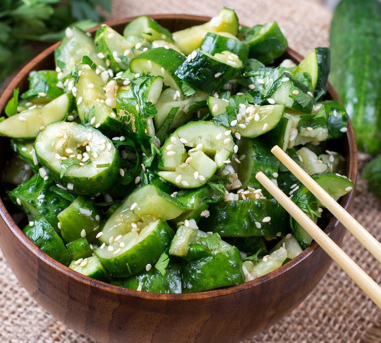 Eine brauen Schüssel mit dem Gurkensalat auf einem hellgrauen Untergrund mit Gurken und Koriander. | © Shutterstock/ Ahanov Michael