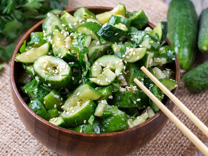 Eine brauen Schüssel mit dem Gurkensalat auf einem hellgrauen Untergrund mit Gurken und Koriander. | © Shutterstock/ Ahanov Michael