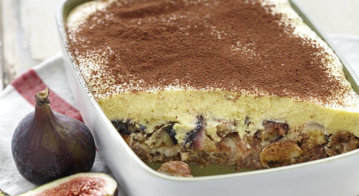 Eine Auflaufform voller Feigen-Tiramisu, angeschnitten, sodass die Feigen unter der Mascarponecreme zu sehen ist. Serviert auf einem hellen Holztisch und garniert mit Kakaopulver. 