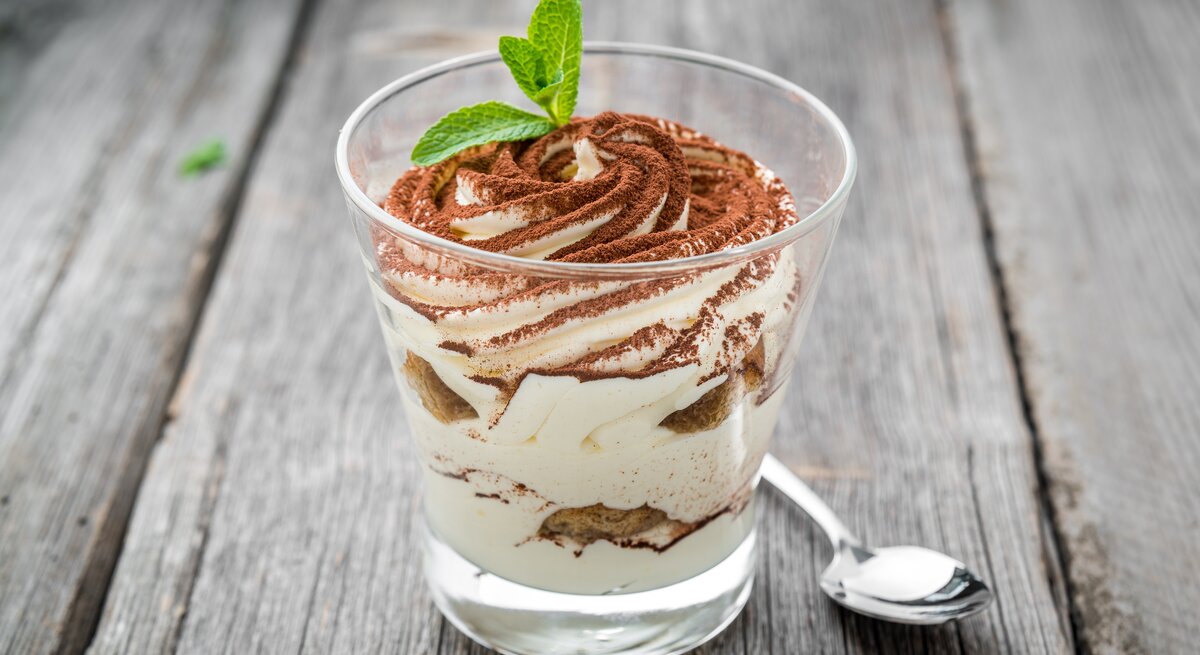 Ein durchsichtiges Glas gefüllt mit einem Dessert bestehend aus gezuckerter Kondensmilch (Milchmädchen) und Gebäck. Das Ganze steht auf einem grauen Holztisch. 