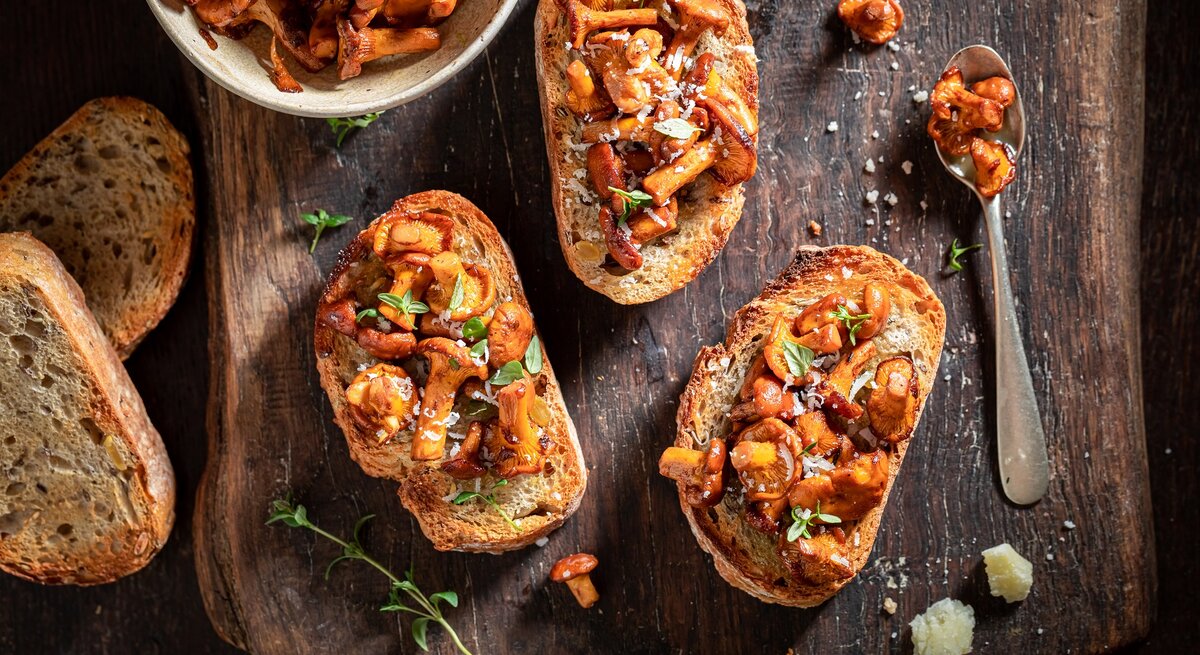 Selbstgemachte Knoblauch-Bruschetta mit Pfifferlingen liegen auf einem Untergrund aus Holz. Daneben liegen ein Löffel mit Pilzen, einige Brotscheiben und eine Schüssel mit Pilzen.