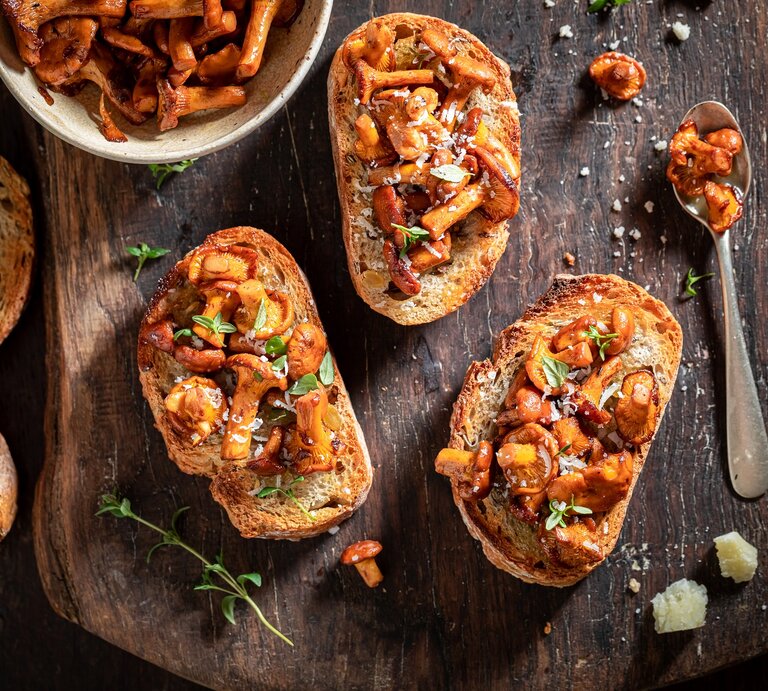Selbstgemachte Knoblauch-Bruschetta mit Pfifferlingen liegen auf einem Untergrund aus Holz. Daneben liegen ein Löffel mit Pilzen, einige Brotscheiben und eine Schüssel mit Pilzen. | © Shutterstock / Shaiith