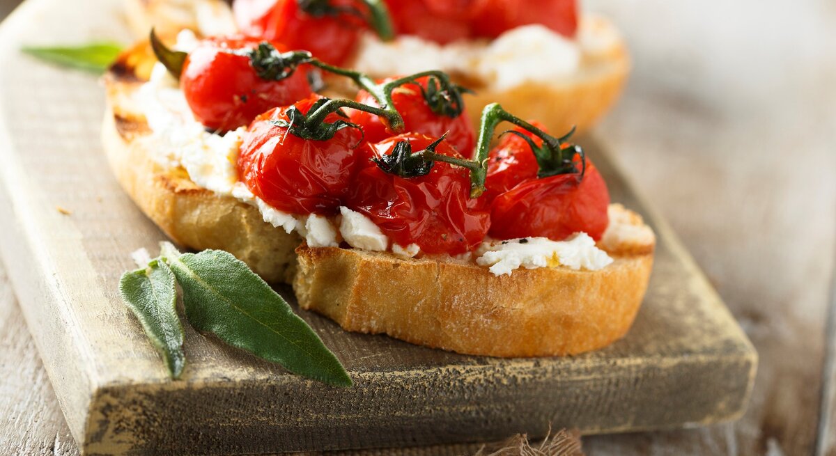Gegrillte Bruschetta mit geschmolzenen Tomaten und Salbei auf hellem Holzbrett und grauer Stoffserviette. Im Hintergrund unscharf eine weitere. Frontalansicht.