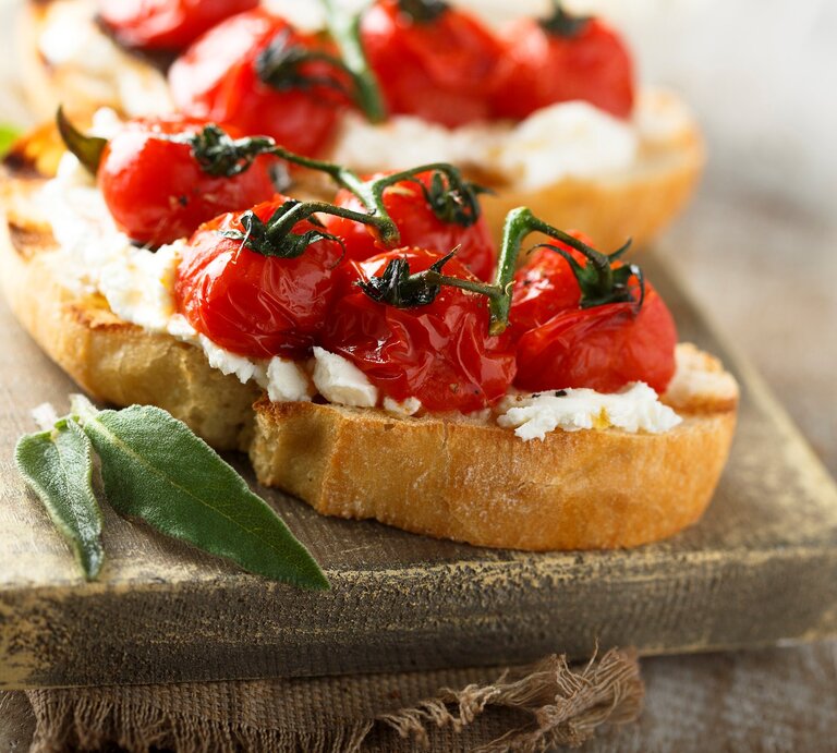Gegrillte Bruschetta mit geschmolzenen Tomaten und Salbei auf hellem Holzbrett und grauer Stoffserviette. Im Hintergrund unscharf eine weitere. Frontalansicht. | © Getty Images / Mariha-kitchen