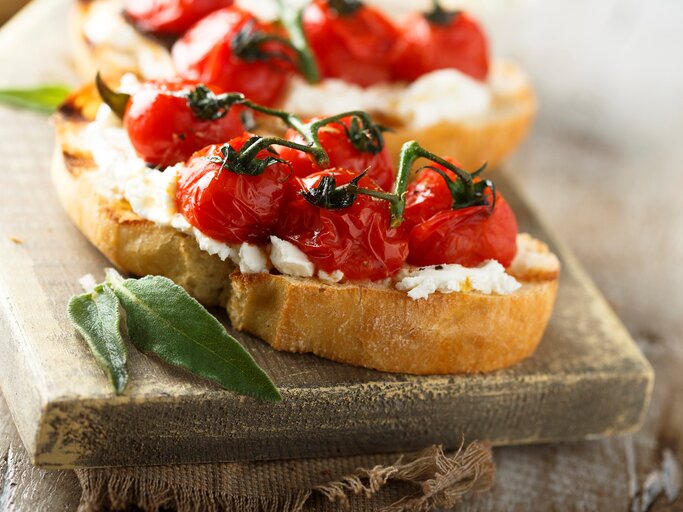 Gegrillte Bruschetta mit geschmolzenen Tomaten und Salbei auf hellem Holzbrett und grauer Stoffserviette. Im Hintergrund unscharf eine weitere. Frontalansicht. | © Getty Images / Mariha-kitchen
