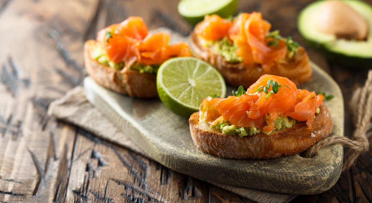 Drei Lachs-Bruschetta mit Avocado auf einem Holztablett.