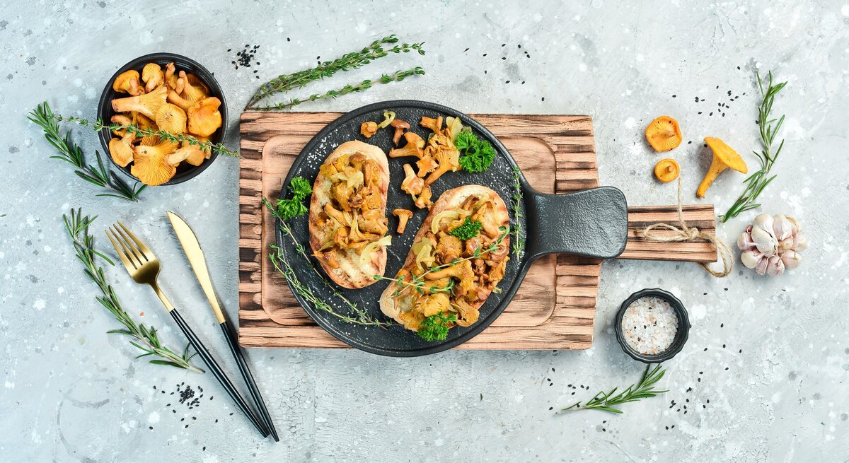 Draufsicht: Heller grau-blauer Untergrund auf dem ein Holzbrettchen mit zwei Käse-Bruschetta mit gebratenen Pfifferlingen legen Ringsherum verstreut sind Knoblauch, Pfifferlinge und Kräuter.