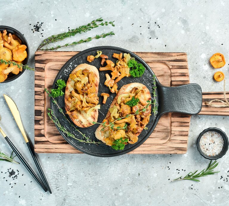 Draufsicht: Heller grau-blauer Untergrund auf dem ein Holzbrettchen mit zwei Käse-Bruschetta mit gebratenen Pfifferlingen legen Ringsherum verstreut sind Knoblauch, Pfifferlinge und Kräuter. | © Shutterstockj / YARUNIV Studio