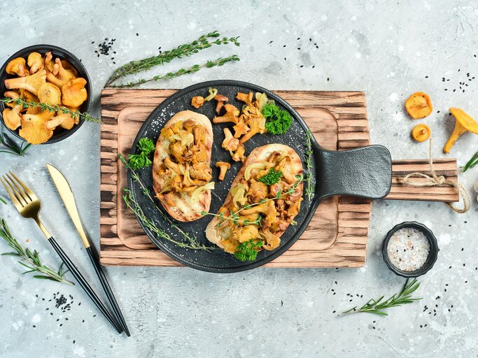 Draufsicht: Heller grau-blauer Untergrund auf dem ein Holzbrettchen mit zwei Käse-Bruschetta mit gebratenen Pfifferlingen legen Ringsherum verstreut sind Knoblauch, Pfifferlinge und Kräuter. | © Shutterstockj / YARUNIV Studio
