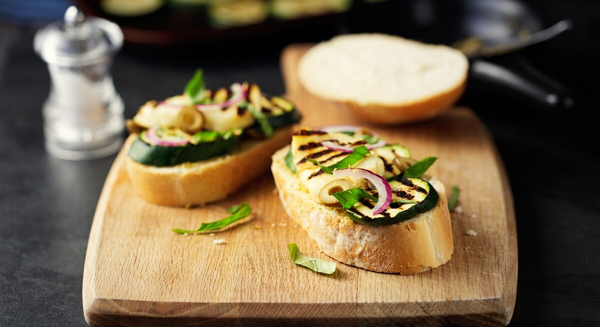 Ein Holzbrett mit Halloumi-Bruschetta mit Zucchini liegt auf einem dunklen Untergrund. Daneben steht ein Salzstreuer.