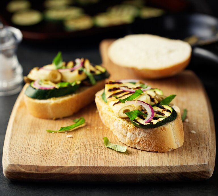 Ein Holzbrett mit Halloumi-Bruschetta mit Zucchini liegt auf einem dunklen Untergrund. Daneben steht ein Salzstreuer. | © Getty Images / haoliang