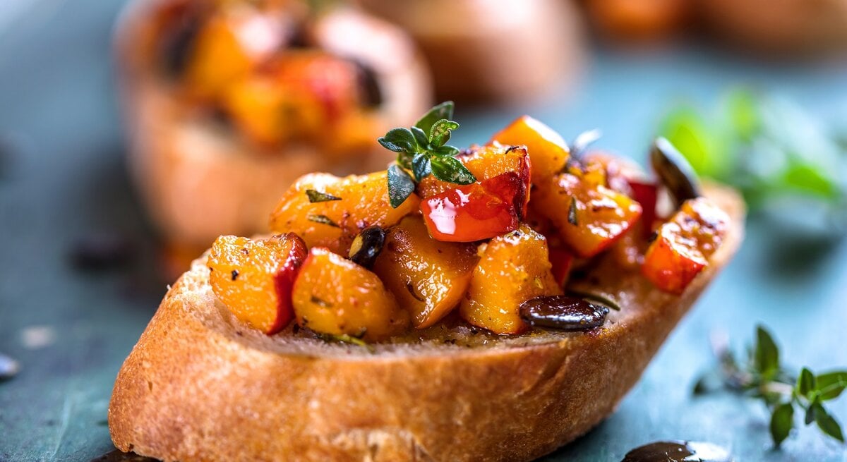Eine Kürbis-Bruschetta mit Kürbiskernen und Kräutern auf einem Stück Baguette, im Hintergrund unscharf weitere Bruschettas.