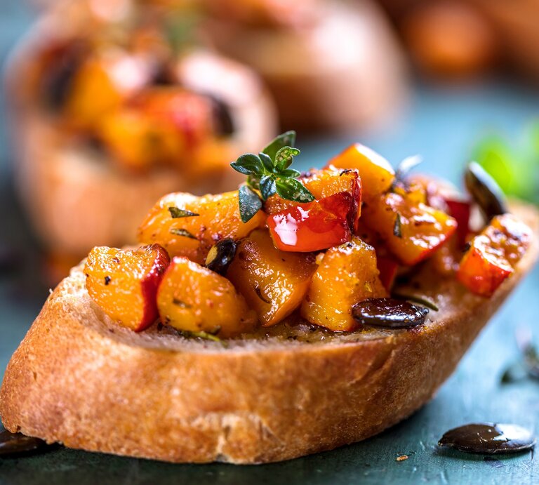 Eine Kürbis-Bruschetta mit Kürbiskernen und Kräutern auf einem Stück Baguette, im Hintergrund unscharf weitere Bruschettas. | © Adobe Stock/ karepa