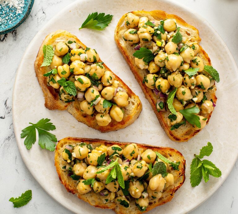 Ein weißer Teller mit drei Scheiben Bruschetta auf einem Marmoruntergrund direkt von oben fotografiert. Daneben ein weiß-braunes Tuch und etwas Salz und Olivenöl. | © Shutterstock/ Elena Veselova