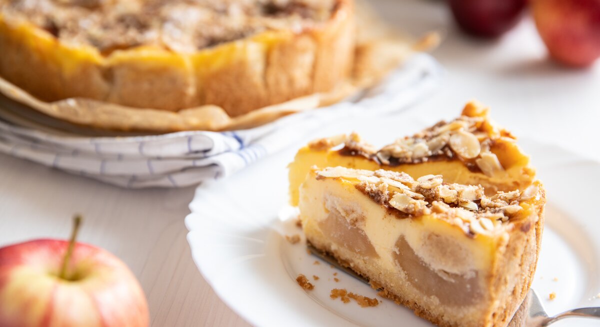 Angeschnittener Bratapfel-Schmandkuchen mit zwei einzelnen Stücken auf  einem weißen Teller. Der Kuchen präsentiert sich mit einer goldbraunen, mit Mandeln belegten Kruste.