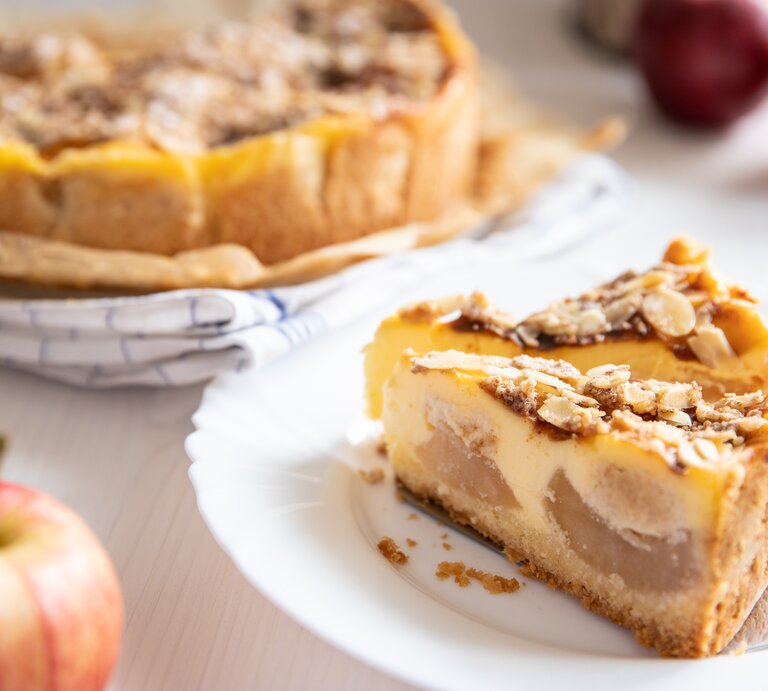 Angeschnittener Bratapfel-Schmandkuchen mit zwei einzelnen Stücken auf  einem weißen Teller. Der Kuchen präsentiert sich mit einer goldbraunen, mit Mandeln belegten Kruste. | © AdobeStock/Kerstin