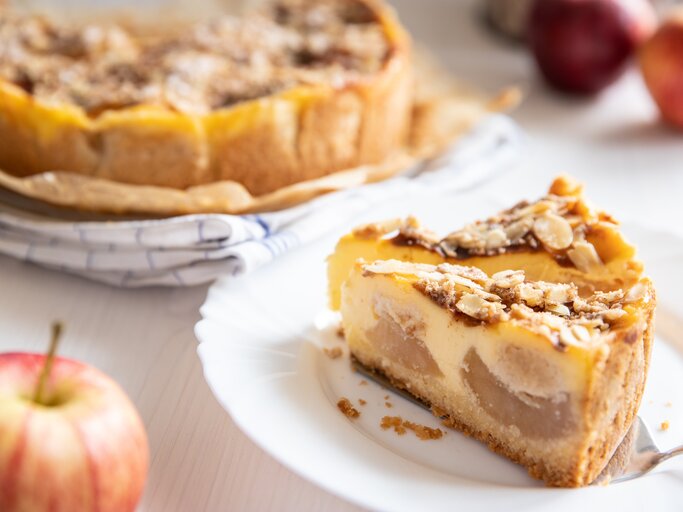 Angeschnittener Bratapfel-Schmandkuchen mit zwei einzelnen Stücken auf  einem weißen Teller. Der Kuchen präsentiert sich mit einer goldbraunen, mit Mandeln belegten Kruste. | © AdobeStock/Kerstin