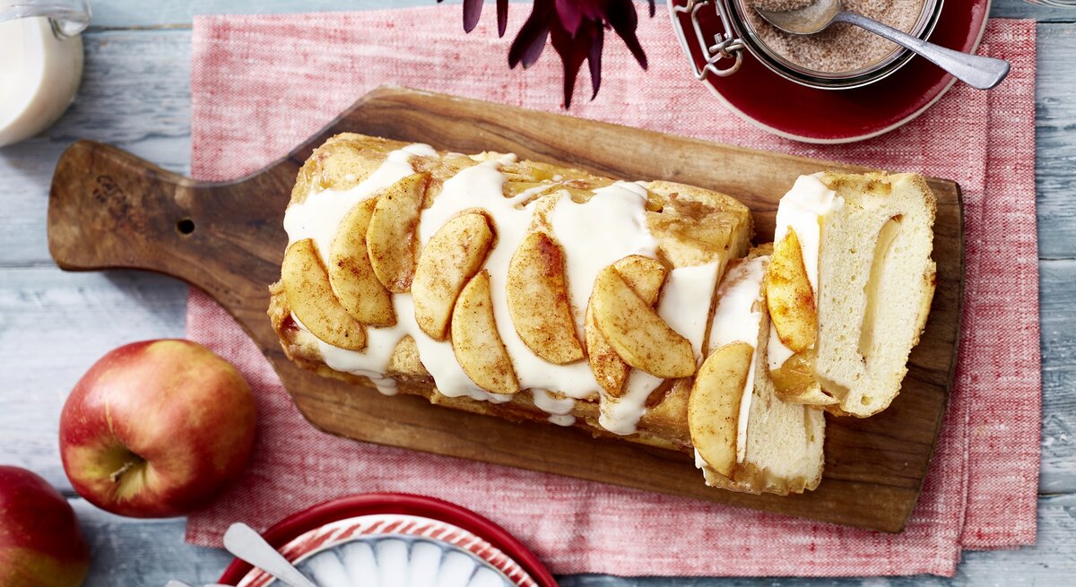 Auf einem Brett liegt ein Apfel-Biskuit-Kuchen aus der Kastenform. Er ist überzogen mit Vanillesoße und mit Äpfeln dekoriert. 