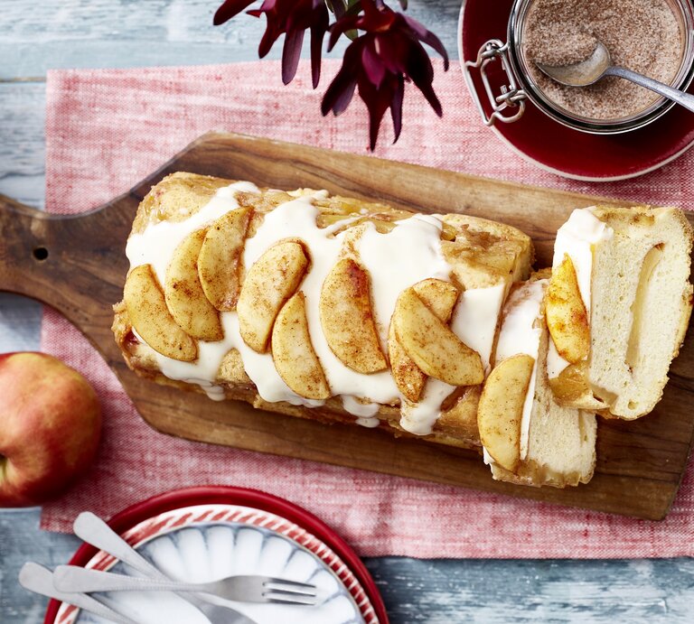 Auf einem Brett liegt ein Apfel-Biskuit-Kuchen aus der Kastenform. Er ist überzogen mit Vanillesoße und mit Äpfeln dekoriert.  | © EAT CLUB/Isabell Triemer