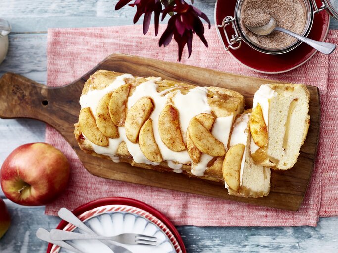 Auf einem Brett liegt ein Apfel-Biskuit-Kuchen aus der Kastenform. Er ist überzogen mit Vanillesoße und mit Äpfeln dekoriert.  | © EAT CLUB/Isabell Triemer