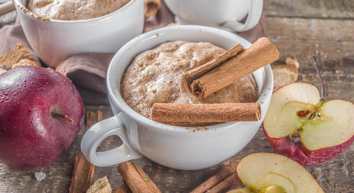 Zwei Tassen mit Apfel-Zimt-Tassenkuchen neben frischen Äpfeln auf einem Holztisch von schräg oben fotografiert.
