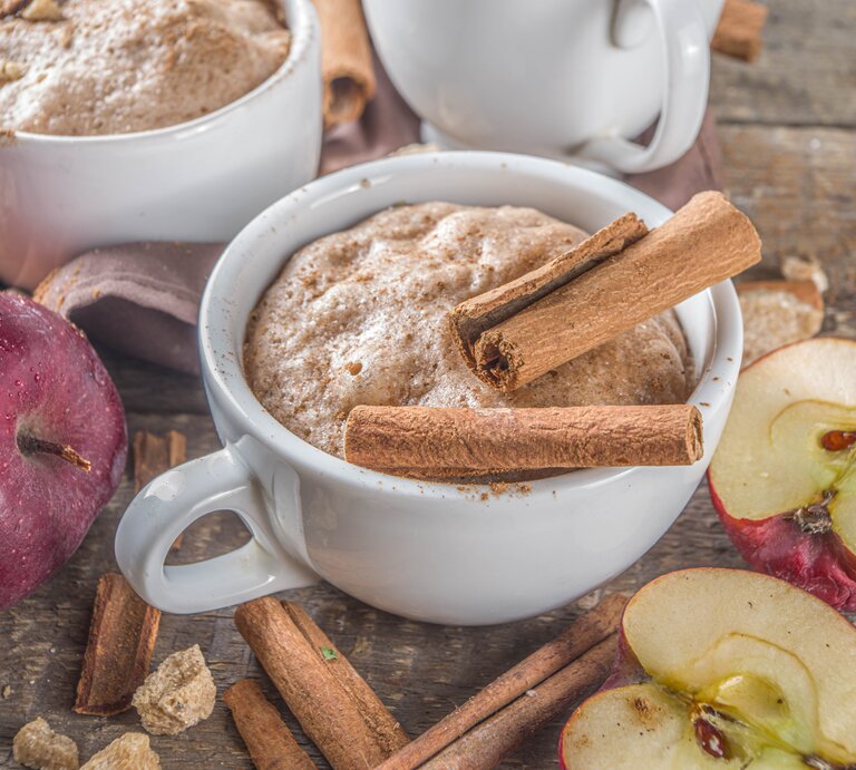 Zwei Tassen mit Apfel-Zimt-Tassenkuchen neben frischen Äpfeln auf einem Holztisch von schräg oben fotografiert. | © Shutterstock/Rimma Bondarenko