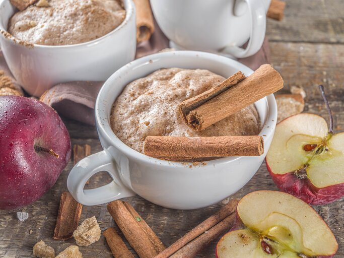 Zwei Tassen mit Apfel-Zimt-Tassenkuchen neben frischen Äpfeln auf einem Holztisch von schräg oben fotografiert. | © Shutterstock/Rimma Bondarenko