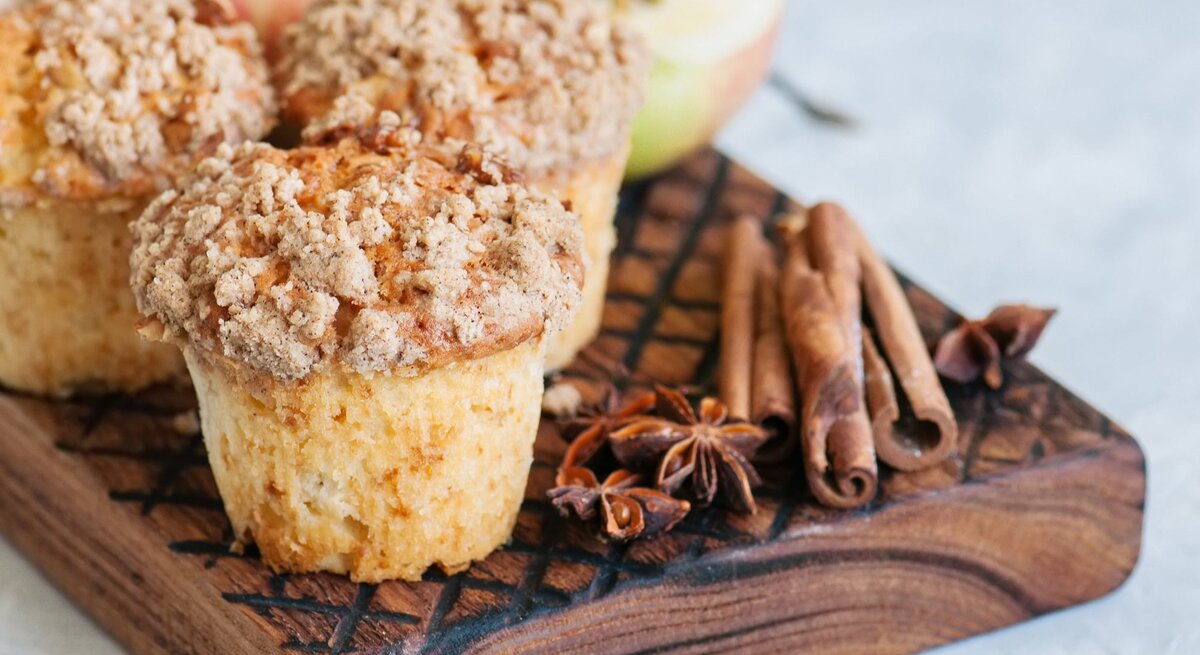 Drei Apfel-Streusel-Muffins auf einem Brett mit Anissternen und Zimtstangen, im Hintergrund unscharf halbierte Äpfel.